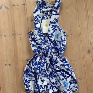 Byxdress  - Blå blommig playsuit från WINONA! Kommer med tillhörande snörning. Snygg urringning framtill. Stl S