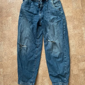 Blå jeans från Zara - Säljer ett par blå jeans från Zara med slitna detaljer på knäna. De har en klassisk femficksdesign och knappgylf. Perfekta för en avslappnad look.