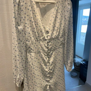 Vit blommig klänning från Nelly x Glamorous - Säljer en söt vit klänning med blått blommönster från Nelly. Klänningen har en v-ringad design och knappar framtill.  Slutar ovanför knäna🌸använd en gång.