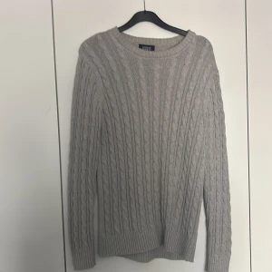 Grå stickad tröja från Iconic Sport - Säljer en stilren grå stickad tröja från Iconic Sport. Tröjan har ett klassiskt flätmönster och långa ärmar, perfekt för kyligare dagar. Den är mjuk och bekväm, idealisk för både vardag och avslappnade tillfällen.