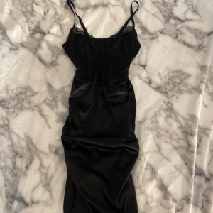 Svart klänning med spetsdetaljer - Elegant svart klänning med smala axelband och spetsdetaljer vid bysten. Tillverkad i ett glansigt material som ger en lyxig känsla. Perfekt för en kväll ute eller en speciell tillställning.