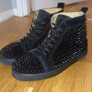 !!Äkta!! Christian Louboutin -  !!Äkta!!Snygga svarta sneakers i mocka från Christian Louboutin med  nitar över hela ytan. Skorna har den ikoniska röda sulan och snörning framtill. Perfekta för att ge din outfit en edgy touch.