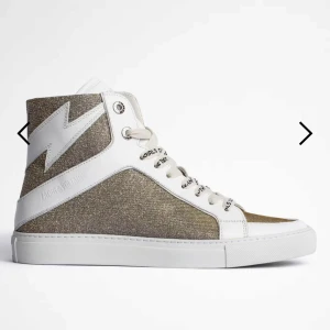 Glittriga sneakers från Zadig & Voltaire - Snygga sneakers med glittrig guld och vit design från Zadig & Voltaire. Skorna har en hög modell med snörning och en cool blixtdetalj på sidan. Perfekta för att ge din outfit en extra touch av stil.