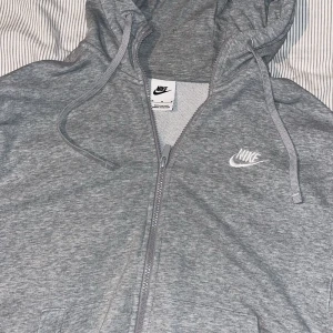 Grå hoodie från Nike/ Zipper - Säljer en stilren grå hoodie från Nike med dragkedja och huva. Perfekt för en avslappnad look. Den har en broderad Nike-logga på bröstet och är tillverkad i mjukt material för extra komfort. Inte sliten alls precis som ny