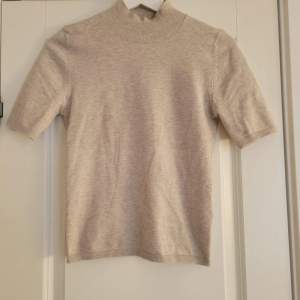 Säljer en stilren beige tröja med korta ärmar och hög krage. Perfekt för en minimalistisk look. Tröjan har ribbade kanter vid ärmslut och nederkant.