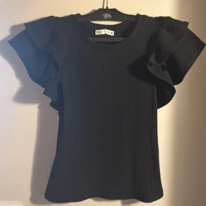 Svart ribbad topp från Zara - Snygg och söt svart ribbad topp från Zara med volangdetaljer på ärmarna. Endast använd 1 eller 2 gånger tror jag. Strl S men Xs funkar säkert också!