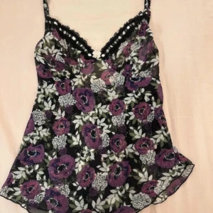 Blommig linne med spetsdetaljer - Säljer en blommig linne med lila och vita blommor på en svart bakgrund. Toppen har smala axelband och spetsdetaljer vid halsringningen. Perfekt för en somrig look!