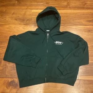 Mörkgrön hoodie med tryck - Säljer en mörkgrön hoodie med dragkedja och tryck både fram och bak. Den har en klassisk passform med långa ärmar och en bekväm huva. Perfekt för en avslappnad stil.