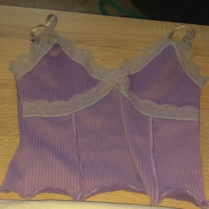 Lila ribbad linnetopp från H&M Divided - Säljer en söt lila ribbad linnetopp från H&M Divided. Toppen har spetsdetaljer längs kanterna och justerbara axelband. Perfekt för en avslappnad stil eller att bära under en kofta.