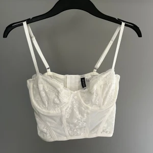 Vit spetskorsett från H&M Divided - Säljer en elegant vit korsett från H&M Divided med vacker spetsdetalj. Den har justerbara axelband och hakknäppning bak. Perfekt för att ge en stilfull touch till din outfit.