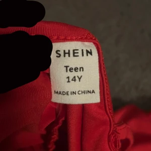 Röd ärmlös klänning från Shein - Snygg röd ärmlös klänning från Shein i storlek 14Y. Klänningen har en åtsittande passform med rynkade detaljer längs sidorna och mitten, vilket ger en cool look. Perfekt för att sticka ut med sin starka färg och stilrena design.