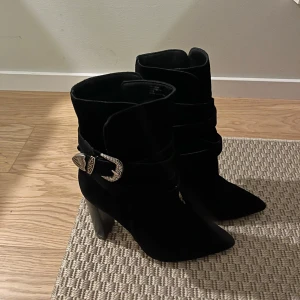 Svarta boots med spänne - Snygga svarta boots i mocka med ett dekorativt spänne i silver. De har en spetsig tå och en stabil klack som ger en elegant touch. Perfekta för att lyfta vilken outfit som helst.