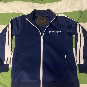 Blå träningsjacka från Palm Angels - Snygg blå träningsjacka från Palm Angels med vita ränder längs ärmarna och dragkedja framtill. Jackan har en sportig look med ribbade muddar och två fickor med dragkedja. Perfekt för en casual stil eller träning.
