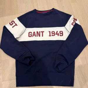 Mörkblå sweatshirt från GANT - Säljer en mörkblå sweatshirt från GANT med vit panel och röd text 'GANT 1949' på bröstet. Tröjan har långa ärmar och ribbade muddar. Perfekt för en avslappnad stil.