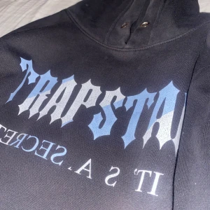 Svart hoodie med trapstar märke - Säljer en svart hoodie med ett stort tryck i blått och vitt på framsidan. Hoodien har en klassisk design med känguruficka och långa ärmar. Perfekt för en avslappnad stil. Vid snabb köp kan priset diskuteras 