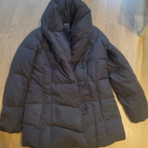 Svart dunjacka från Esprit - Säljer en svart dunjacka från Esprit med stora knappar framtill. Jackan har en pösig krage och är perfekt för kyliga dagar. Den är långärmad och har en stilren design som passar till det mesta. Perfekt för dig som vill hålla värmen med stil!