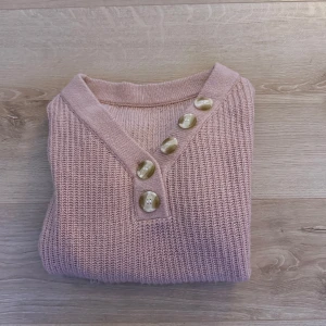 Rosa stickad tröja med knappar - Säljer en söt rosa stickad tröja med v-ringning och dekorativa knappar framtill. Perfekt för en mysig dag. Tröjan har långa ärmar och en ribbad kant vid ärmslut och nederkant.