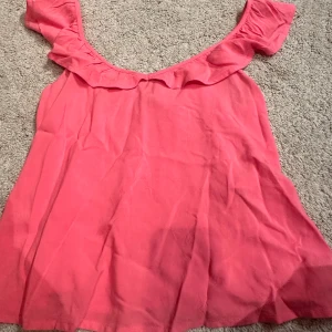 Rosa topp med volangdetalj - Säljer en söt rosa topp med volangdetalj vid halsringningen. Toppen har korta ärmar och en luftig passform, perfekt för varma dagar. Den är gjord i ett mjukt och bekvämt material.