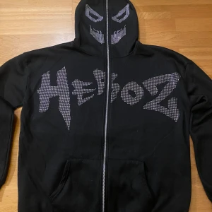 Svart huvtröja med strassdetaljer - Cool svart huvtröja med dragkedja och strassdetaljer. Framsidan har texten 'HeBoZ' i strass och huvan har ett ansiktsmönster. Baksidan pryds av strassvingar. Perfekt för en edgy look! Mycket bra skick! Storlek M