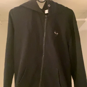 Svart hoodie med dragkedja från Play - Säljer en svart hoodie med dragkedja från Play. Den har ett litet hjärta med ögon på bröstet och en stor ögonapplikation på ryggen. Perfekt för en casual look.