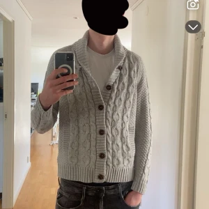 Grå stickad kofta med knappar - Säljer en stilren grå stickad kofta med knappar. Koftan har ett snyggt flätmönster och en bred krage som ger en extra touch. Perfekt för kyligare dagar. Tröjan passar personer mellan 187-193cm 