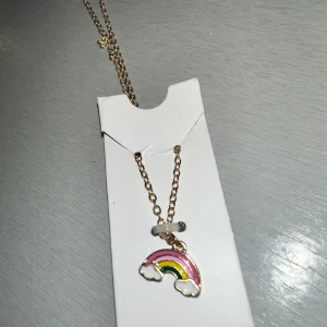 Halsband med regnbågshänge - Charmigt halsband med ett färgglatt regnbågshänge. Hänget har rosa, gul och grön färg med vita moln på sidorna. Kedjan är i guldton och har en enkel design som passar till hänget.