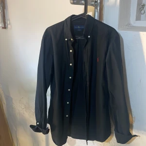 Svart skjorta från Ralph Lauren - Snygg svart skjorta från Ralph Lauren med klassisk krage och knappar framtill. Den har en röd broderad logga på bröstet som ger en stilren touch. Perfekt för en elegant look.