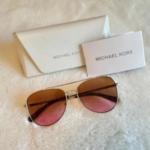 Michael Kors solglasögon MK 1045 San Diego - Snygga Michael Kors solglasögon, modell MK 1045 San Diego. De har en stilren design med guldiga bågar och tonade glas i brunt och rosa. Perfekta för en solig dag med en touch av lyx.