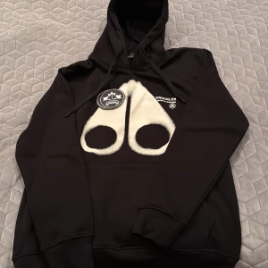 Svart hoodie från Moose Knuckles - Säljer en svart hoodie från Moose Knuckles med en unik vit design på framsidan. Hoodien har en justerbar huva och är perfekt för kyliga dagar. Märket är känt för sin kvalitet och stil. Passar bra för en avslappnad look.