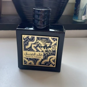 Qaed Al Fursan Eau de Parfum - Elegant parfymflaska i svart med guldmönster och en springande häst på etiketten. Flaskan har en fyrkantig form och en matchande svart kork. Parfymen är tillverkad i UAE och är en Eau de Parfum. Ca 90% kvar.