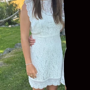 Vit spetsklänning - Säljer en söt vit spetsklänning med fina detaljer. Klänningen är ärmlös och har en elegant spetsdesign med knäppning i nacken. Perfekt för sommarens alla tillfällen.