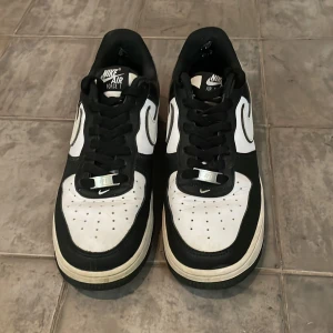 Nike Air Force 1 i svart och vitt - Snygga Nike Air Force 1 sneakers i klassisk svart och vit färgkombination. Skorna har en vit bas med svarta överlägg och den ikoniska Nike-loggan på sidan. Perfekta för en stilren look.