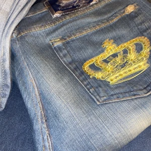 Blå jeans med guldbroderad krona - Snygga blå jeans med en unik guldbroderad krona på bakfickan. Perfekt för dig som vill sticka ut med en kunglig touch. Jeansen har en klassisk design med dragkedja och knapp framtill.