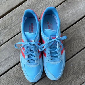 Adidas LA Trainer blå/röda sneakers - Snygga Adidas LA Trainer sneakers i ljusblå mocka och textil med röda detaljer och klassiska tre ränder på sidorna. Skorna har snörning, platt sula och rund tå. Perfekta för dig som gillar retrostil och vill sticka ut med färg.