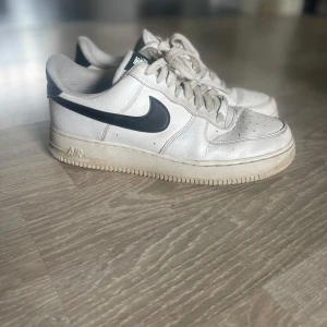 Nike Air Force 1 vita sneakers - Klassiska Nike Air Force 1 sneakers i vitt skinn med svart swoosh och detaljer. Lågt skaft, perforerad tå och snörning framtill. 