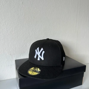Svart New Era NY Yankees keps 7 1/8 - Klassisk svart New Era 59FIFTY keps med New York Yankees-logga broderad i vitt framtill. Rak skärm och broderad MLB-logga baktill. Tillverkad i polyester för en clean och stilren look. Perfekt för dig som vill ha streetstyle-vibe.