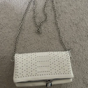 Vit axelväska med nitar från Even&Odd - Snygg vit axelväska från Even&Odd med silverfärgade nitar i mönster på framsidan. Väskan har en kedjerem i metall och dragkedja framtill. Perfekt storlek för det viktigaste och ger en cool vibe till din outfit.