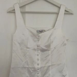 Vit satinlinnetopp från NA-KD - Supersnygg vit linnetopp i satin från NA-KD med dekorativa knappar framtill. Toppen har breda axelband och figurnära passform. Aldrig använd och helt ny med prislapp.