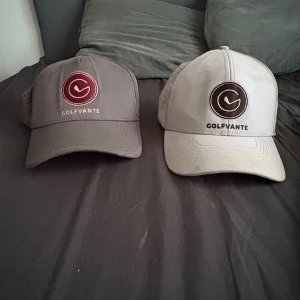 Två kepsar från Golfvante med mesh - Säljer två snygga kepsar från Golfvante, en i mörkgrått med vinröd logga och en i ljusgrått med svart logga. Båda har böjd skärm, ventilerande meshpaneler med hål och justerbar snapback baktill. Perfekta för golf eller andra utomhusaktiviteter.