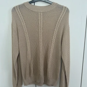 Beige stickad tröja från Lindex - Säljer en beige stickad tröja från Lindex i storlek 170. Tröjan har snygga flätstickade detaljer framtill och ribbade muddar vid ärmslut och nederkant. Perfekt för dig som gillar en enkel men ändå stilren look. Aldrig använd
