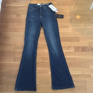 Blå bootcut jeans från NA-KD - Snygga mörkblå bootcut jeans från NA-KD med hög midja och klassisk femficksdesign. Jeansen har en lätt tvättad look och är tillverkade i stretchig denim för en bekväm passform. Perfekta för dig som gillar retrovibbar och markerade ben. 