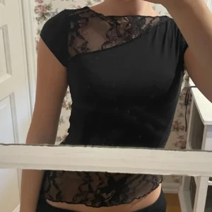 Svart topp med spetsdetaljer - Snygg svart topp med transparent spets upptill och nertill. Asymmetrisk design med korta ärmar och croppad passform. Spetsen har blommigt mönster som ger en cool och feminin vibe. Perfekt för dig som vill sticka ut lite extra.