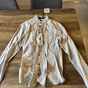 Ljusgrå skjorta från Blend, storlek S - Säljer en stilren ljusgrå skjorta från Blend i storlek S. Skjortan har klassisk krage, bröstficka och svarta knappar hela vägen. Tillverkad i mjuk bomull och passar perfekt till jeans eller chinos. Enkel och clean look för dig som gillar minimalistisk stil.