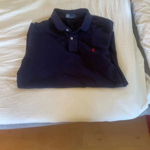Mörkblå pikétröja Polo Ralph Lauren storlek m - Säljer en klassisk mörkblå pikétröja från Polo Ralph Lauren i storlek M. Tröjan har korta ärmar, krage med två knappar och den ikoniska röda logotypen broderad på bröstet. Perfekt för en clean och stilren look.