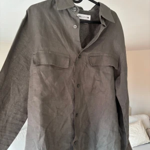 Olivgrön linneskjorta från Zara - Snygg olivgrön skjorta från Zara i 100% linne. Skjortan har klassisk krage, knäppning framtill och två bröstfickor med lock. Perfekt för en chill och stilren look, passar lika bra öppen som stängd. Stryks till innan den skickas!