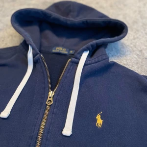  Ralph Lauren Hoodie - Snygg mörkblå hoodie från Polo Ralph Lauren med dragkedja, vita snören och klassisk gul broderad logga på bröstet. Tröjan har huva, känguruficka och är tillverkad i mjuk bomull. Perfekt för en chill och stilren look.