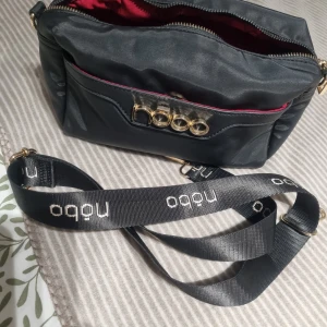 Svart axelväska från Nobo med gulddetaljer - Snygg svart axelväska från Nobo med bred axelrem med logga och guldfärgade metalldetaljer framtill. Väskan har dragkedja upptill och röd insida som ger en cool kontrast. Perfekt för att bära dina essentials med stil.