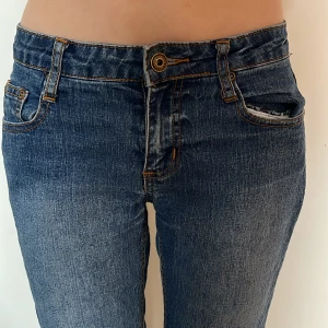 Blå Lågmidjade bootcut jeans med broderad ficka - Snygga blå jeans med bootcut-snitt och låg midja. Jeansen har klassisk femficksdesign, orange kontrastsömmar och en broderad bakficka med vågigt mönster. Tillverkade i jeansmaterial med lätt tvättad look. Perfekta för en avslappnad stil. 