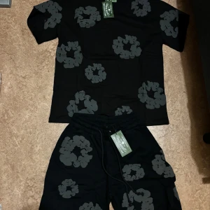 Denim tears tracksuit/ set - Svart t-shirt från Denim Tears med gråa blommiga tryck över hela plagget. Klassisk passform med rund hals och korta ärmar. Tillverkad i mjuk bomull, perfekt för en chill och trendig look.