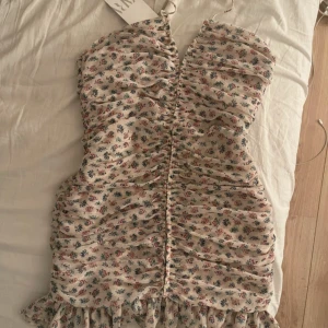 Blommig kortklänning från Zara - Supersöt kortklänning från Zara med blommigt mönster i rosa, blått och grönt på ljus botten. Klänningen har volangkant nertill, rynkade detaljer och v-ringning. Perfekt för sommarens alla tillfällen. Materialet känns lätt och luftigt.